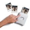White Instax mini printer with three Polaroid-style photos popping out as a hand presses the circular button on the device.
