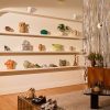 Boutique showroom with white shelves displaying pottery, a plant, and handbags, plus a wooden cabinet and metallic curtain on the right.