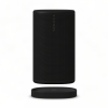 Black cylindrical Sonos wireless speaker with perforated grille, standing next to its round charging dock on a white background.
