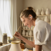 Woman in a white shirt paints a ceramic vase with a small brush in a bright pottery studio with shelves of pottery in the background