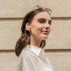 Portrait of a woman in a white shirt with sunglasses perched on her head, facing slightly to the right against a beige wall.