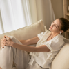 Woman relaxing on a beige sofa with eyes closed, wearing a white blouse and wireless earbuds.