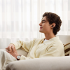 Man sitting on a light sofa with eyes closed, wearing wireless earphones and a cream sweater, relaxing.