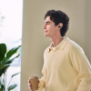 Profile view of a young man wearing a wireless earbud and a pale yellow polo, holding a takeaway coffee cup indoors.