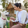 Woman in white top and light jacket sits on grass, smiling as a dog with a pink collar offers its paw in a park.