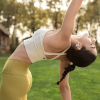 Woman in a white sports bra and yellow leggings performing a backbend yoga pose on a sunny lawn, wearing wireless earphones.