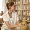 Woman in a white shirt shaping a clay vase on a pottery wheel in a studio with shelves of pottery in the background.