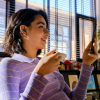 Woman in a lavender striped sweater smiles at her smartphone while holding a coffee cup and wearing a wireless earbud in a bright cafe.