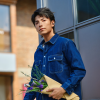 Young man in a denim shirt holds a bouquet wrapped in brown paper outdoors by a building.