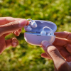 People hand a white wireless earbud case open outdoors, showing earbuds inside against a green blurred background.