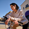 Woman leaning on a beige bicycle outdoors on a sunny day, wearing a light zip-neck sweatshirt and wireless earbuds.