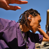 Portrait of a person with braided hair wearing a white wireless earbud, outdoors on a sunny day in a purple shirt and chains.