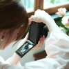 Person holds a black DSLR with the LCD screen flipped out, framing a shot indoors near a window with flowers nearby.