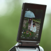 Camera LCD showing a woman and a child under a blue umbrella in a flower garden, being framed by the photographer's viewfinder.