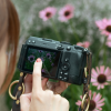 Person holding a DSLR-style camera up to their eye, finger on the shutter, framing pink flowers seen on the LCD screen.