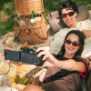 Two friends wearing sunglasses take a selfie at a picnic with a wicker basket, drinks, and snacks on a checkered blanket.