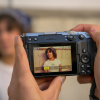 Hands hold a digital camera; on the screen a woman with curly hair poses for a photo in a light background.
