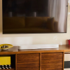 Flat-screen TV mounted above a wooden console with a white Sonos soundbar; a yellow bowl sits to the left and a red container to the right on the cabinet.