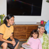 A woman in a yellow shirt sits on the floor with a smiling young girl and another person nearby in a living room setting with a TV and soundbar behind them.