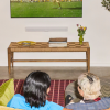 Two people sit on a rug facing a wall-mounted TV with a white soundbar beneath it and a wooden checkerboard coffee table in front, holding a plate of candy, a stack of books, and a vase of tulips.