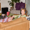 Woman wearing headphones lounges on a brown sofa, smiling at the camera, while a child sleeps beside her under a patchwork quilt.