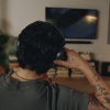 Back view of a person wearing headphones, talking on a phone in a living room with a TV and a stack of books behind them, Tattoos visible on forearm.